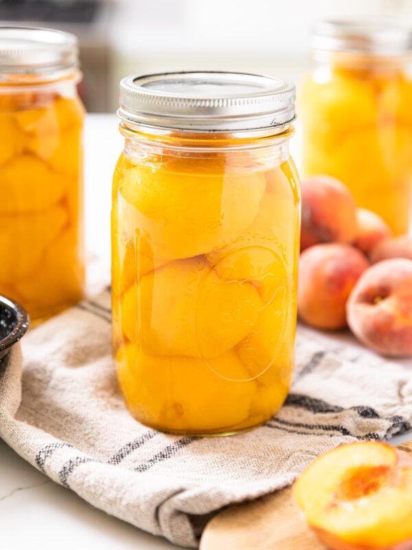 Large glass jar filled with sliced orange colored peaches with clear liquid on top of them with other filled jars in background