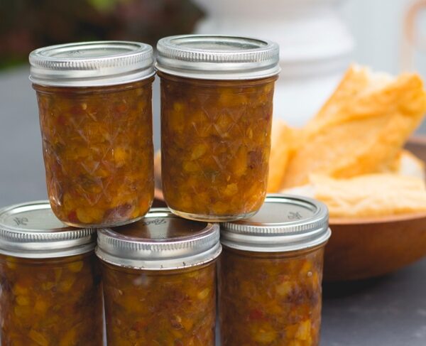 Five canning jars filled with peach chutney with bowl filled with bread in background all on gray slate surface