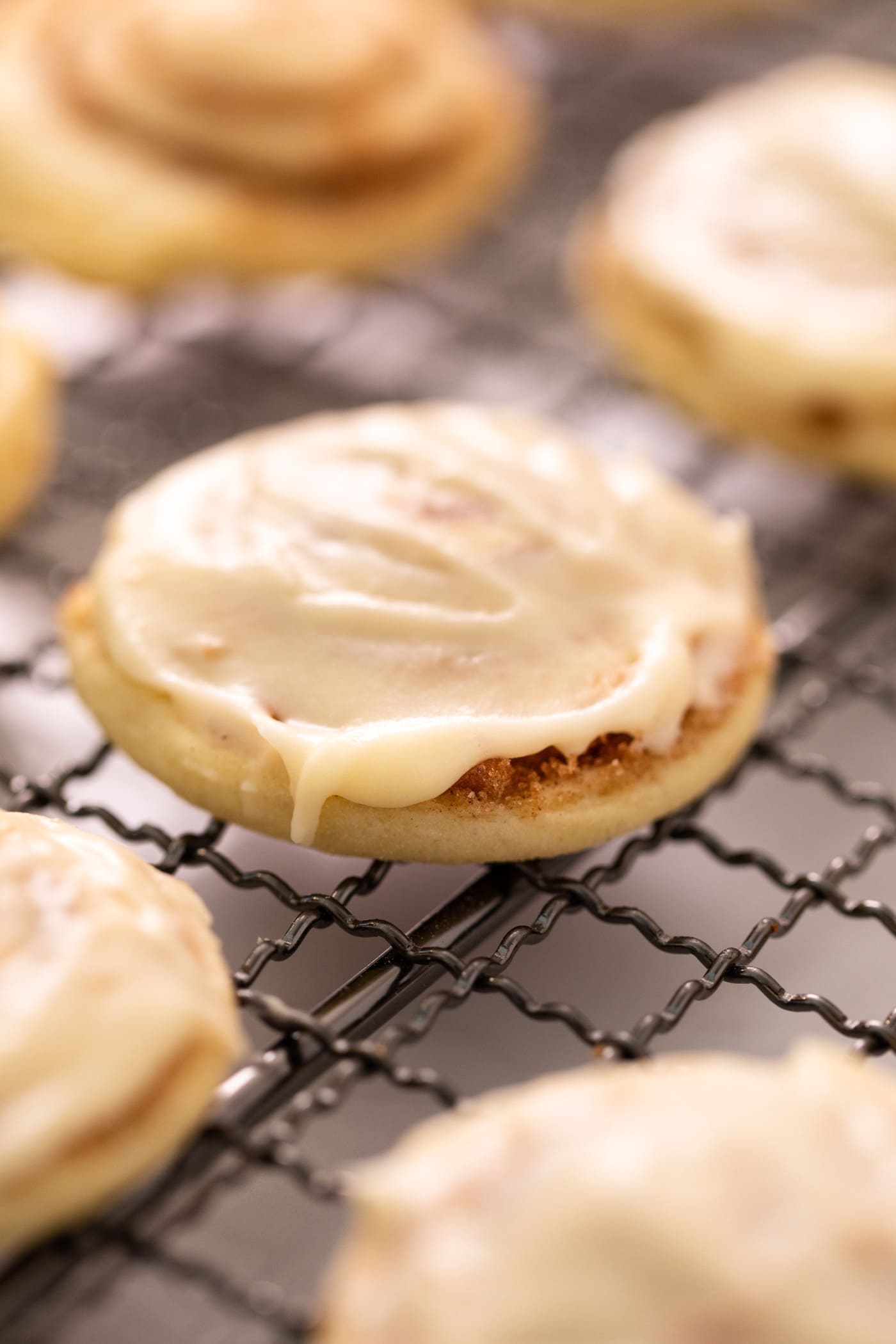 Cinnamon roll cookie with white creamy frosting on top sitting on wire cooling rack with other unfrosted and frosted cookies all around