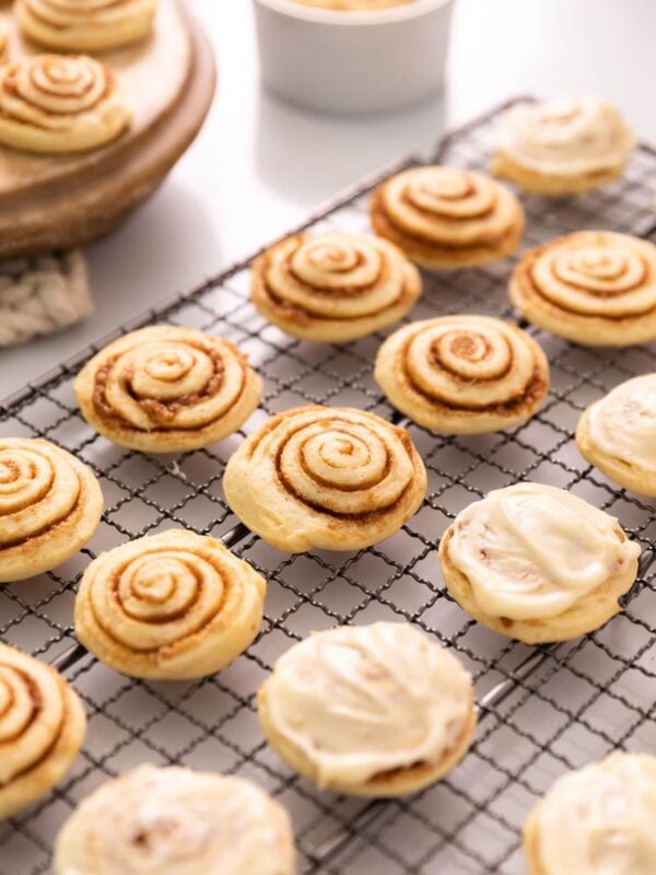 Cinnamon roll cookies with half frosted sitting on wire cooling rack with extra cookies in background along with white container filled with brown sugar