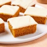 Squares of pumpkin bars with cream cheese frosting all over the top sitting on round white plate.