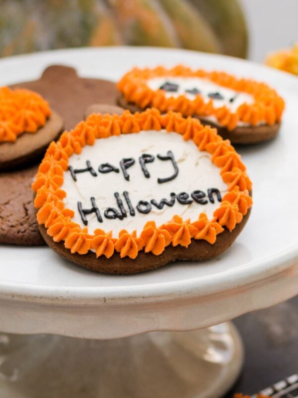 Spiced ginger cutout cookies topped with orange and white frosting sitting on white cake plate with pumpkin in background