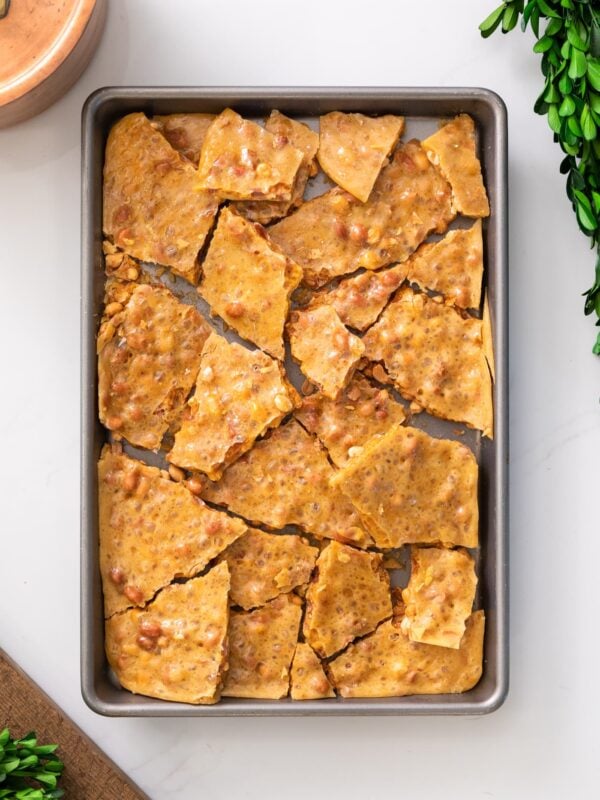 Top down view of shards of yellow colored peanut brittle dotted with peanuts sitting in baking pan all on white marble surface