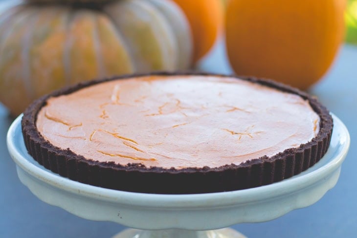 Round cake plate with pumpkin tart with chocolate crust on top with pumpkins in background.