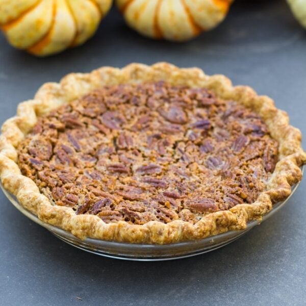 Simple pecan pie topped with pieces of pecans in glass pie dish sitting on gray slate surface with small pumpkins in background
