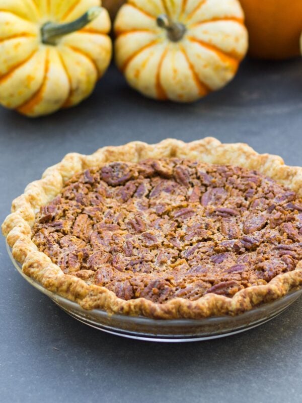 Simple pecan pie in glass pie dish sitting on gray slate surface with small pumpkins sitting in background