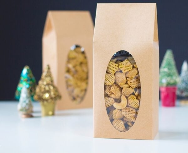 Brown bags filled with caramel nut crispix sitting on white surface with second bag in background with small Christmas trees around.