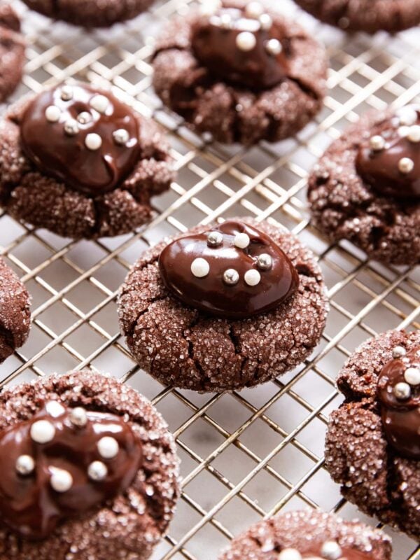 Chocolate cookies with sanding sugar on all edges topped with chocolate ganache and silver and white dragees sitting amongst other cookies on a wire cooling rack.