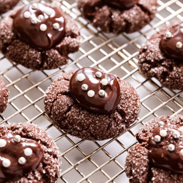 Chocolate cookie with dollop of ganache sitting on top with white and silver dragees on top all on wire cooling rack amongst other chocolate cookies.