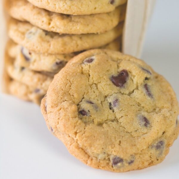 Stack of large chocolate chip cookies sitting on white countertop with one cookie in front and rest of cookies wrapped up in parchment in background