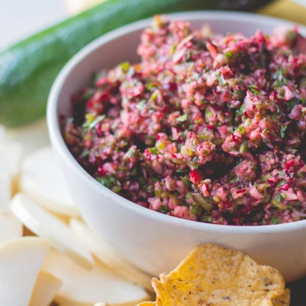 White bowl filled with red colored cranberry salsa with chips and sliced vegetables all around