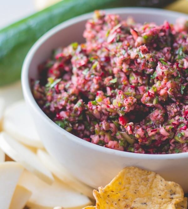 White bowl filled with red colored cranberry salsa with chips and sliced vegetables all around