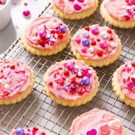 Frosted pink sugar cookies with sprinkles all over the top cooling on a baking rack.