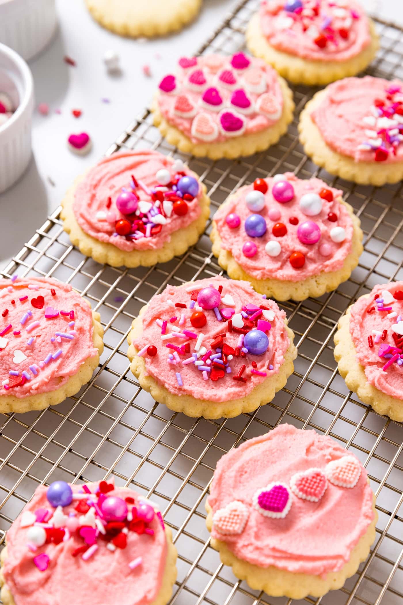 Frosted pink sugar cookies with sprinkles all over the top cooling on a baking rack.