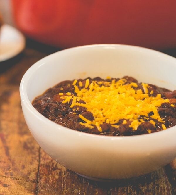 White bowl filled with vegetarian chili topped with sprinkling of cheese sitting on wood surface with red Dutch oven in background