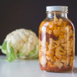 Large half gallon jar sitting on white surface filled with chipotle pickled cauliflower with red peppers in liquid with single head of cauliflower in background