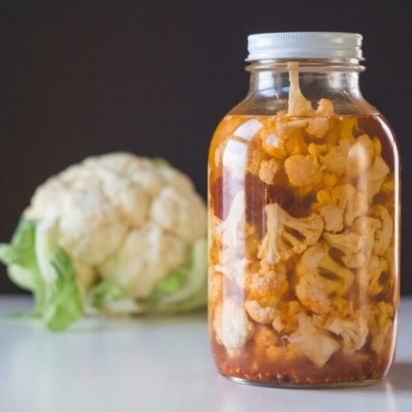 Large half gallon jar sitting on white surface filled with chipotle pickled cauliflower with red peppers in liquid with single head of cauliflower in background