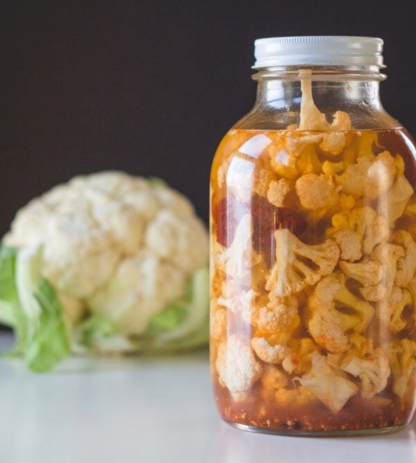 Large half gallon jar sitting on white surface filled with chipotle pickled cauliflower with red peppers in liquid with single head of cauliflower in background