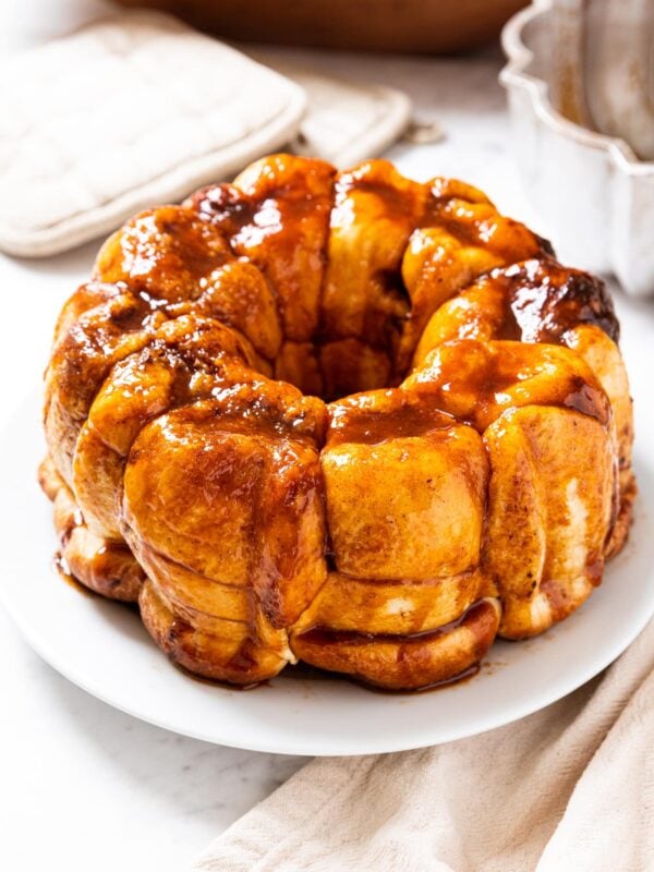 White plate filled with pieces of monkey bread with golden sauce all over the top with glistening edges all on white marble surface with Bundt pan in background.