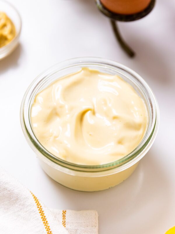 Glass bowl filled with homemade mayonnaise sitting on white countertop with eggs and mustard in background