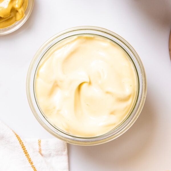 Top down view of glass container filled with homemade mayonnaise sitting on white countertop with lemon and mustard