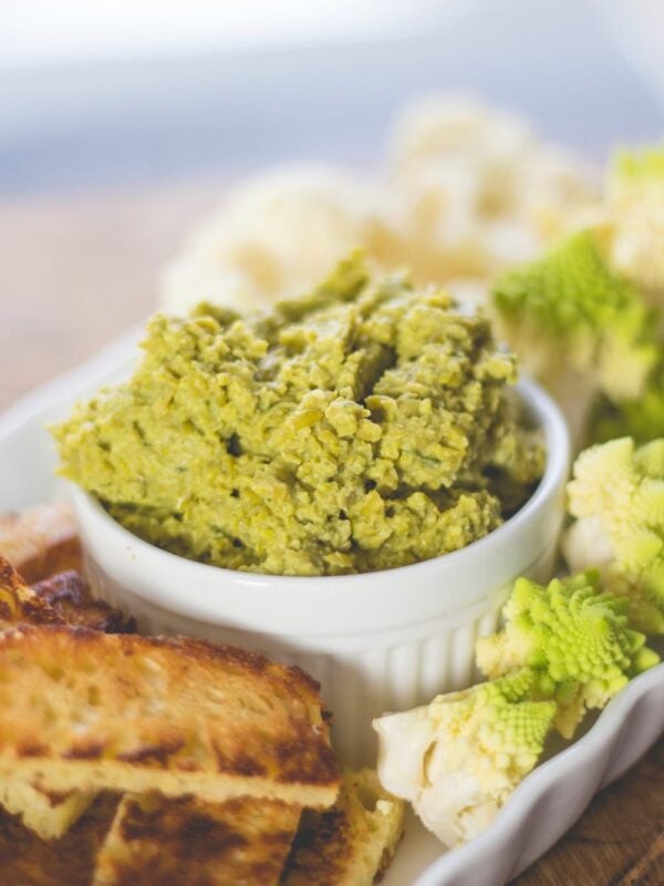 Round white bowl filled with green colored pea hummus sitting on board surrounded by vegetables and slices of crusty bread