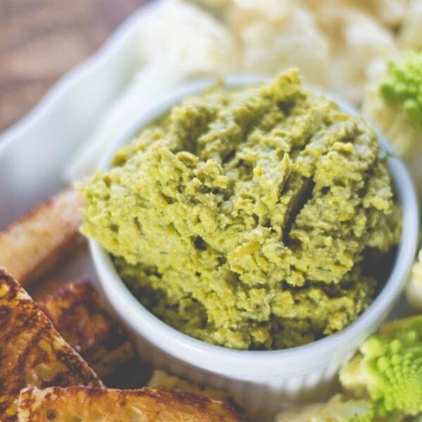 White bowl filled with chunky green-colored pea hummus with sliced vegetables and toasted bread sitting on wood board