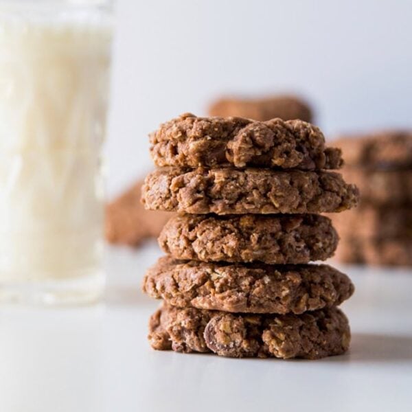 White surface with stack of double chocolate oatmeal cookies with more in the background along with a large glass of milk.