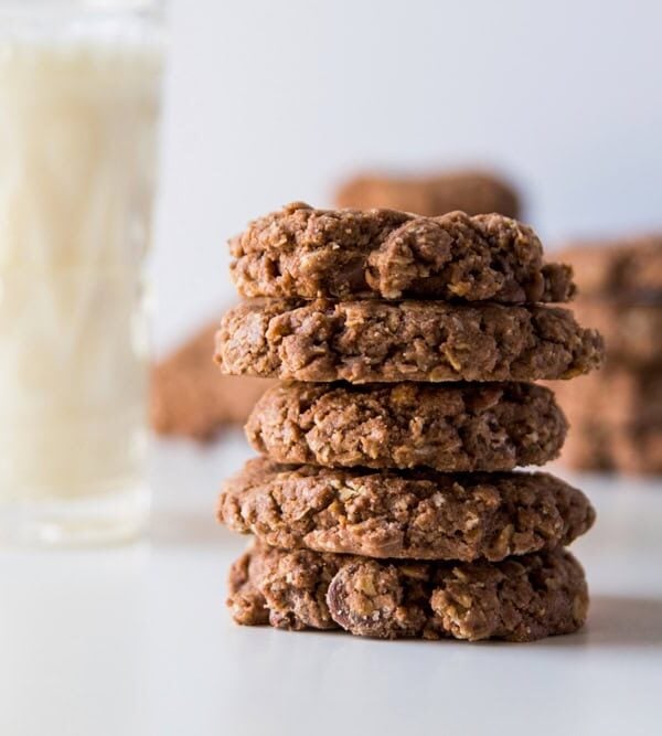White surface with stack of double chocolate oatmeal cookies with more in the background along with a large glass of milk.