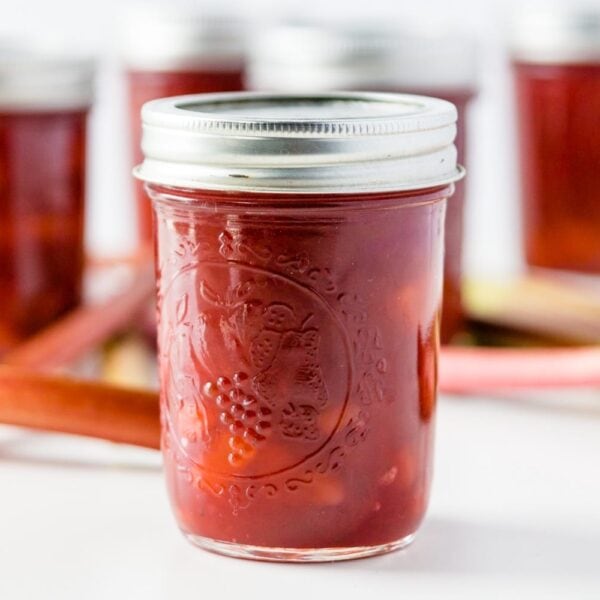 Jar of rhubarb jam with extra jars and rhubarb stalks in background on a white surface