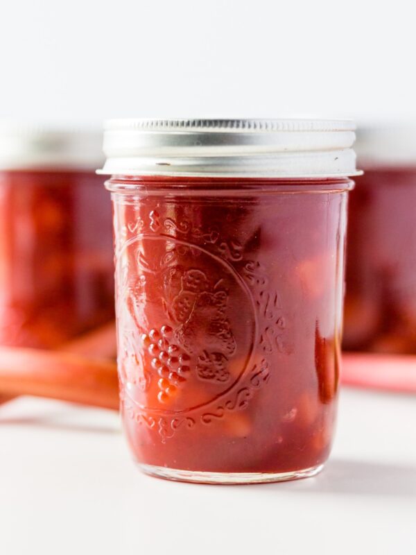 Jar of rhubarb jam with extra jars and rhubarb stalks in background on a white surface