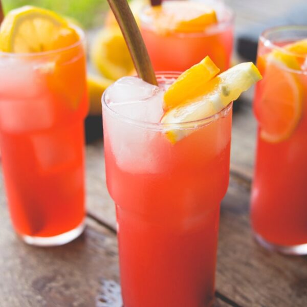 Four glasses filled with bright colored pink rhubarb lemonade with orange slices and stalks of rhubarb in glasses all on wood surface outdoors