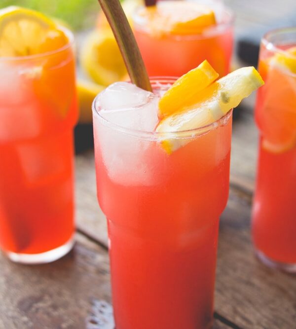 Four glasses filled with bright colored pink rhubarb lemonade with orange slices and stalks of rhubarb in glasses all on wood surface outdoors