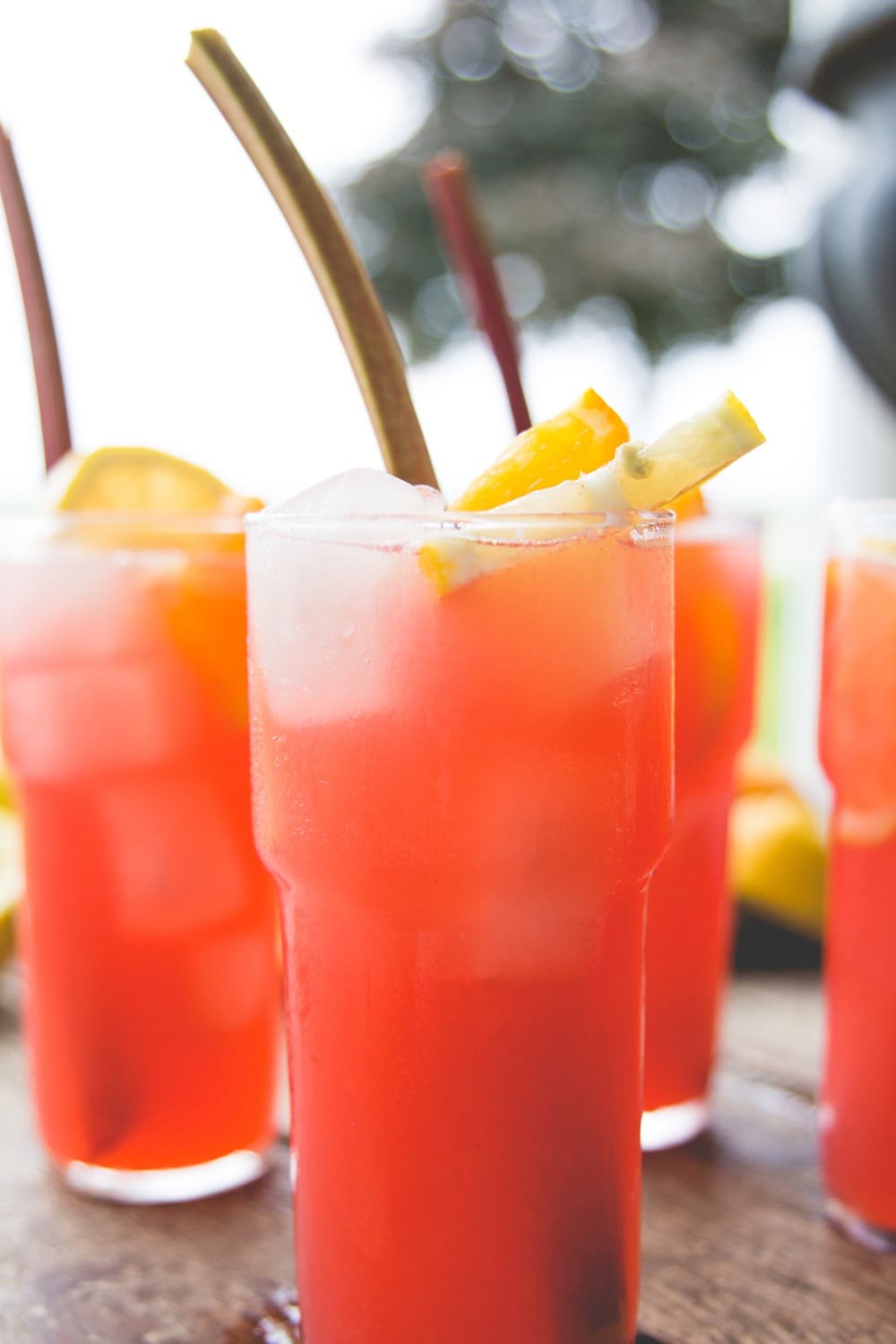 Four large glasses filled with pink colored rhubarb lemonade sitting on wood surface with stalk of rhubarb in glass along with orange slices