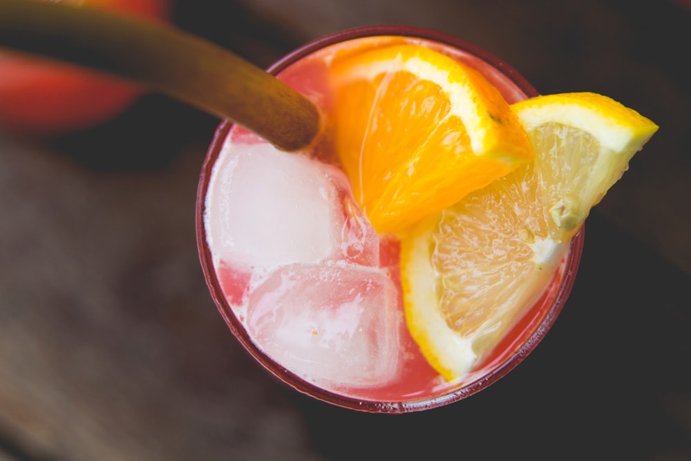 Top down view of glass filled with pink colored rhubarb lemonade with ice cubes, orange and lemon slices and stalk of rhubarb all on wood surface