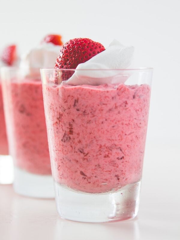 Glasses filled with strawberry rhubarb fool sitting on white surface with slice strawberry and whipped cream