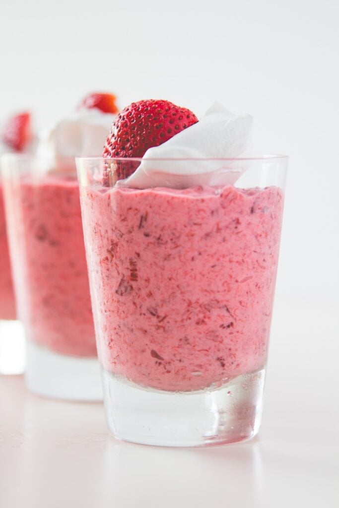 Glasses filled with strawberry rhubarb fool sitting on white surface with slice strawberry and whipped cream