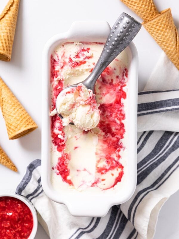 Top down view of white container filled with swirled red and white ice cream with metal ice cream scoop sitting in container with empty ice cream cones all around