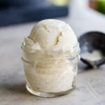Two scoops of vanilla ice cream sitting in glass container with ice scoop and urn in background on marble surface