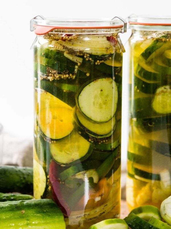 Glass jar of fridge pickles with cucumbers, onions, red peppers and celery seed in light yellow pickling liquid with cutting board and napkin in background