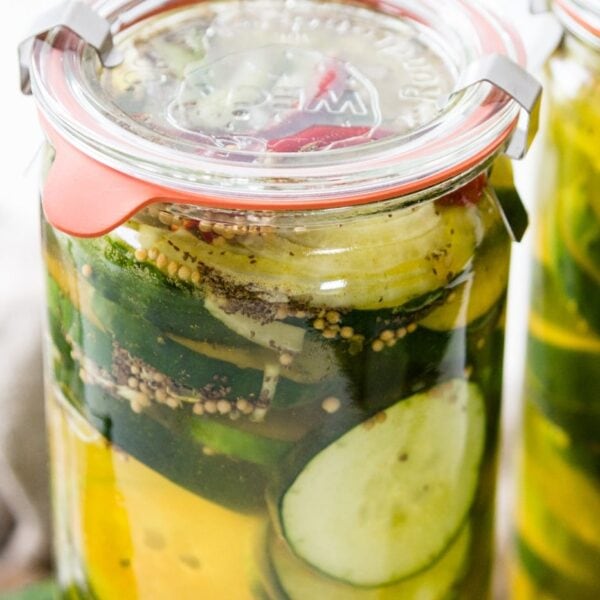 Glass jar of fridge pickles with cucumbers, onions, red peppers and celery seed in light yellow pickling liquid with cutting board and napkin in background