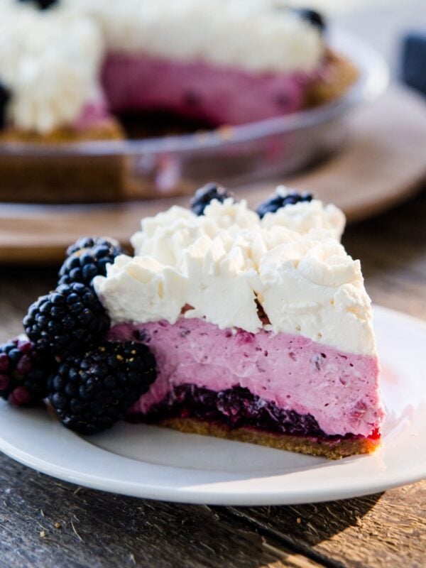 Slice of blackberry icebox pie sitting on white plate with graham cracker crust, blackberry filling and whipped cream layers showing with rest of blackberry pie in background on a wood serving platter