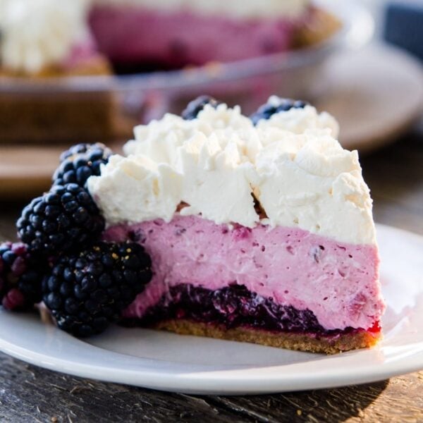 Slice of blackberry icebox pie sitting on white plate with graham cracker crust, blackberry filling and whipped cream layers showing with rest of blackberry pie in background on a wood serving platter