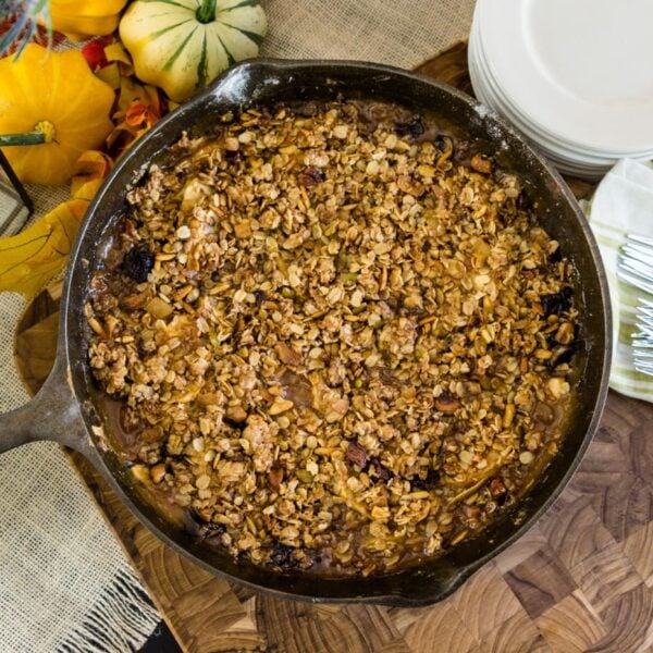 Top down view of skillet apple crisp on a wood cutting board with orange pumpkins and flowers, silverware and plates on a black surface