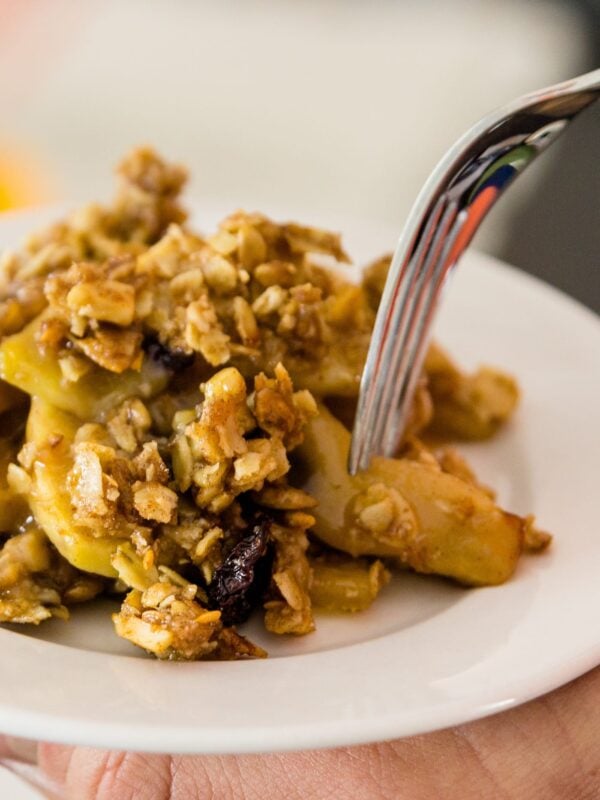 Fork picking up piece of skillet apple crisp sitting on a white plate with hand holding plate
