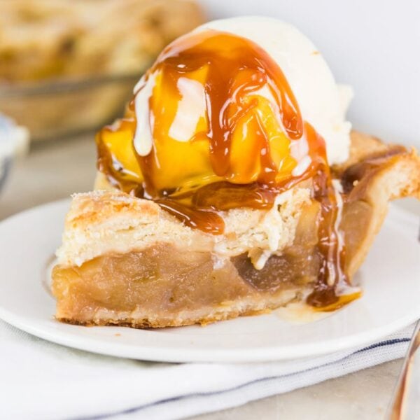 Scoop of ice cream sitting on slice of apple pie with caramel sauce drizzled over top sitting on white plate with napkin
