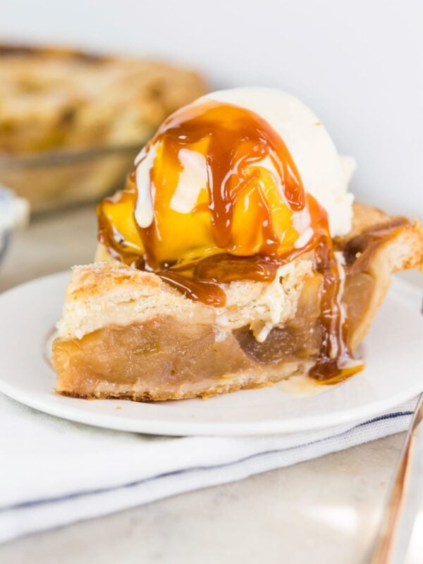 Scoop of ice cream sitting on slice of apple pie with caramel sauce drizzled over top sitting on white plate with napkin