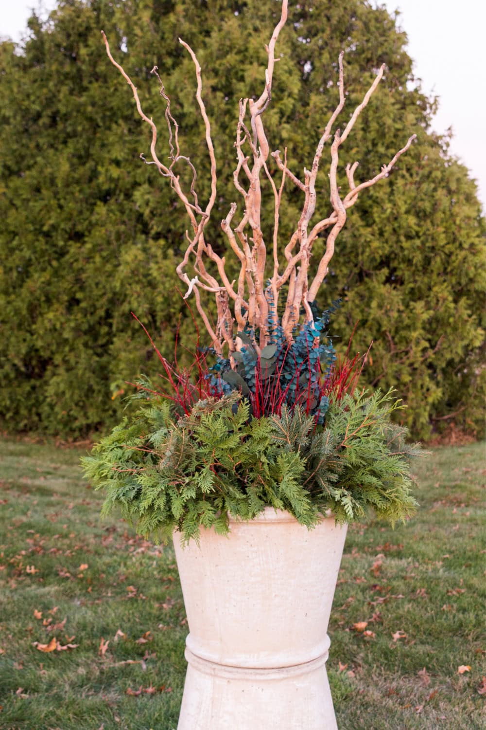 Large concrete urn with greenery around the base with red twigs, eucalyptus, and large branches coming out of the top.