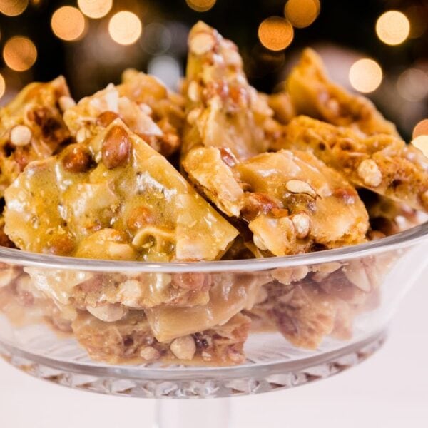 Golden peco candy with peanuts and flakes of coconut piled high in glass bowl on a white surface with Christmas lights in background