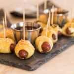 Pigs in a blanket wrapped in dough and sprinkled with sesame seeds all on slate serving platter on marble surface